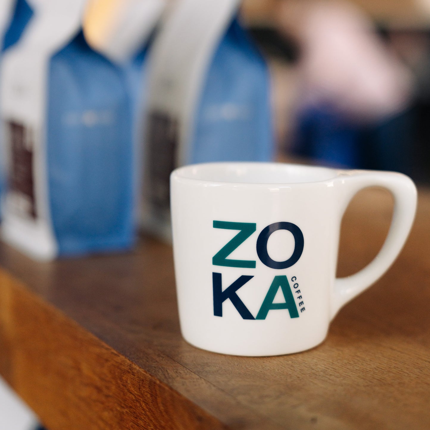 White mug with 'ZOKA' branding on a side, sitting on wooden surface with blurred background