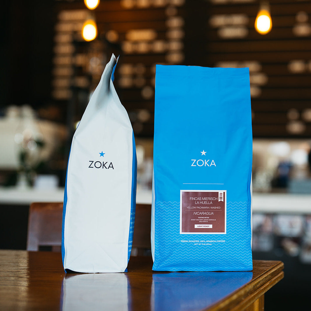 Two Five Pound Zoka coffee bags, one white and one blue, on a wooden surface with a blurred cafe background.