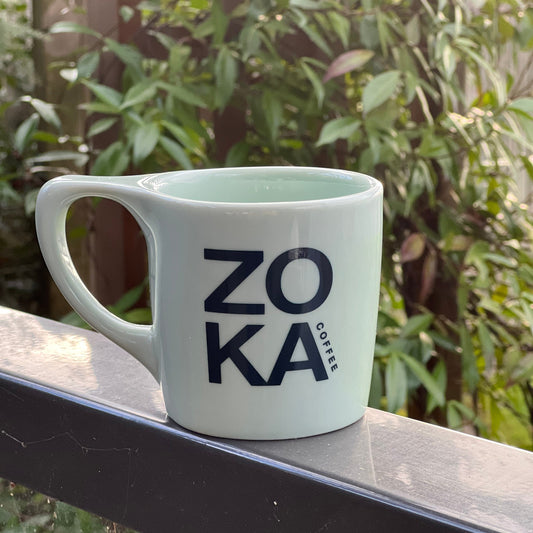 Zonka Coffee mug hanging on a railing with greenery in the background