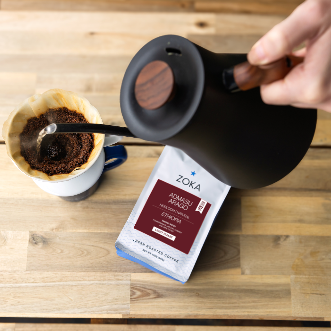 Person pouring coffee from a black kettle into a white mug with a Zoka coffee bag on a wooden surface.