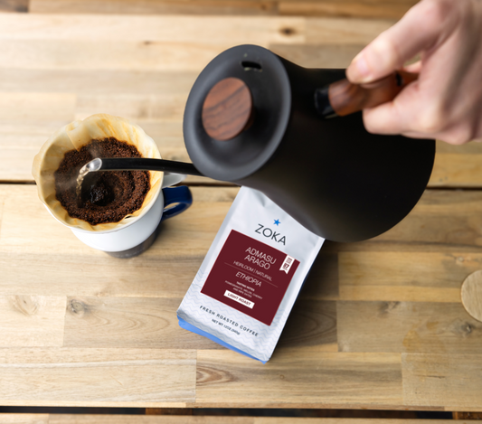 Person pouring coffee from a black kettle into a white mug with a Zoka coffee bag on a wooden surface.