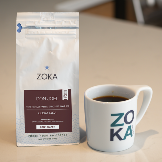 A bag of Zoka Don Joel Costa Rica coffee stands next to a white ceramic mug with the Zoka logo, both on a light beige countertop with a blurred kitchen background.