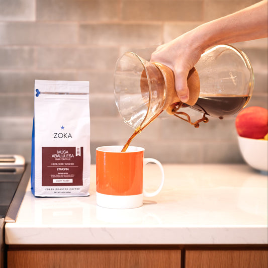 Person pouring coffee from a carafe into an orange mug with Zoka coffee packaging on a kitchen counter.